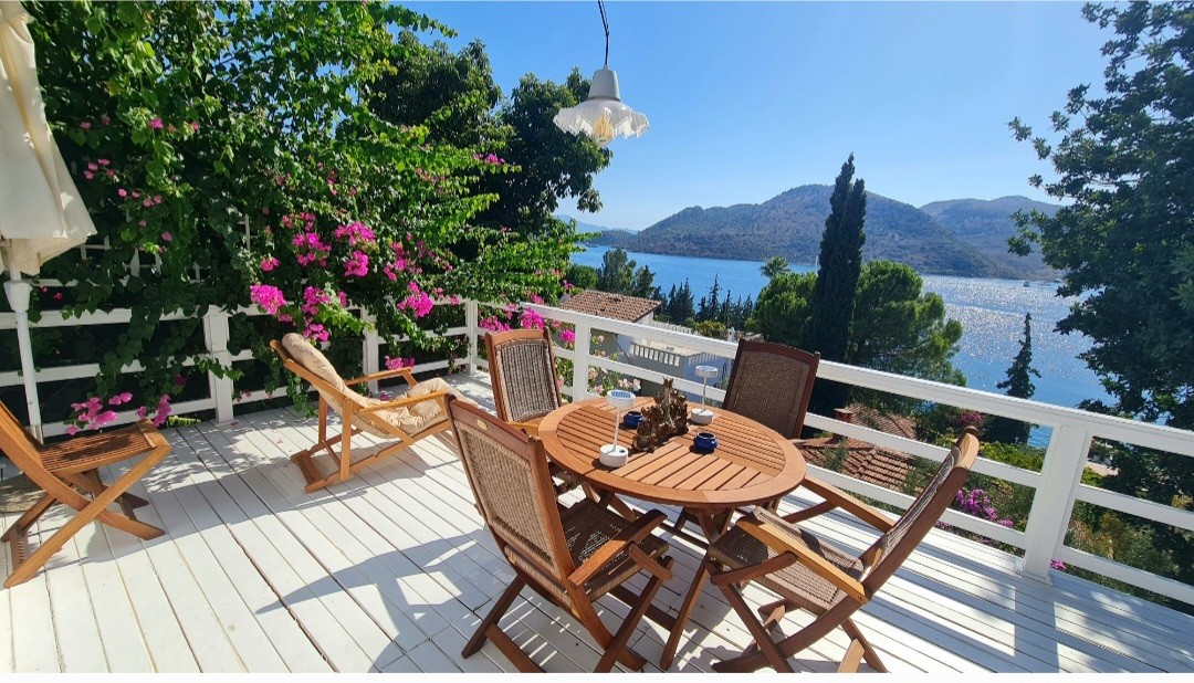 A wooden table with four matching chairs is situated on a sunlit terrace, surrounded by blooming bougainvillea. A scenic view of the turquoise sea and hills can be seen in the background, enhanced by a hanging light fixture above the table.