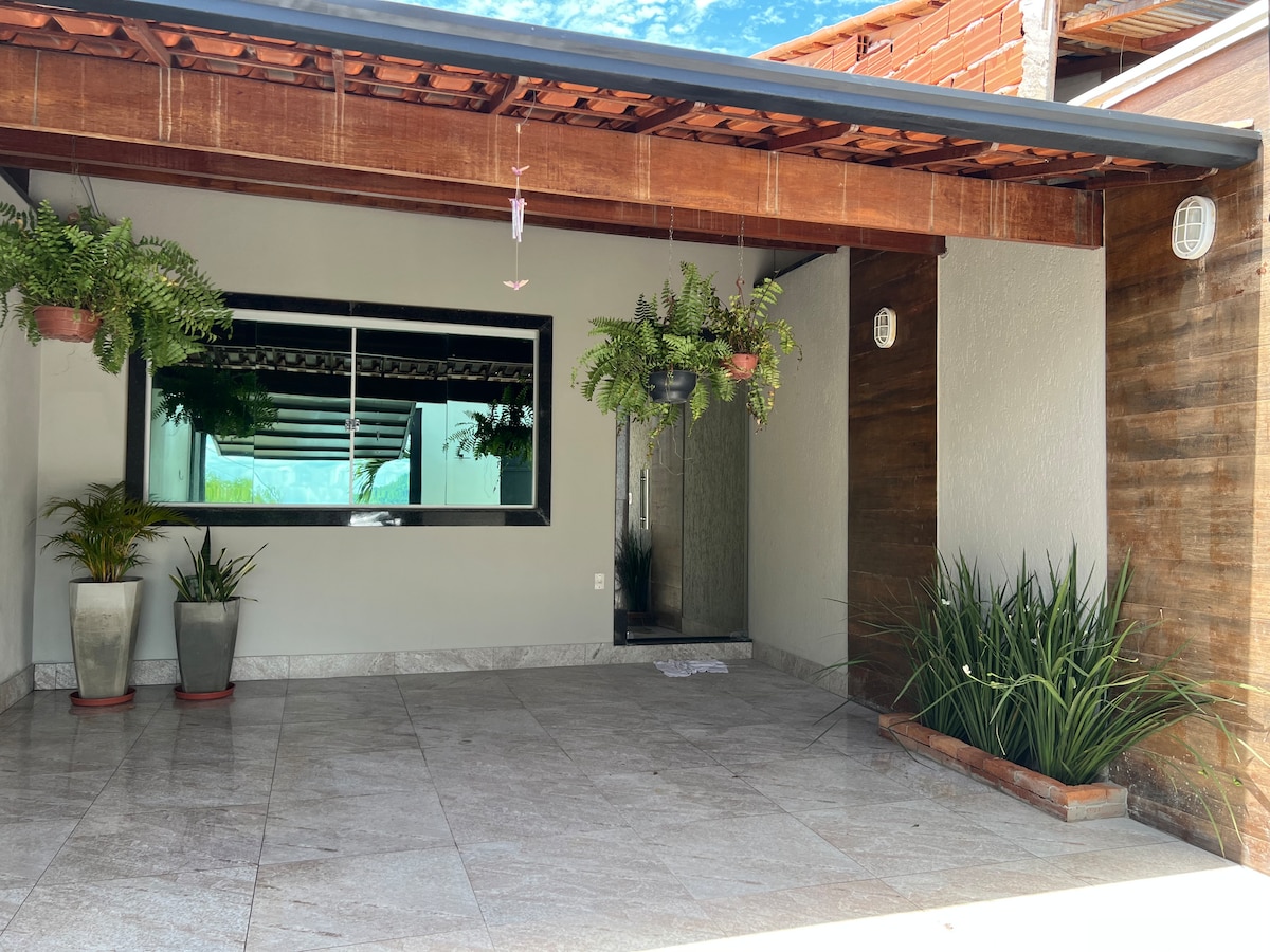 An outdoor area features polished stone flooring and is shaded by a wooden overhang. Large potted plants are positioned on either side, while hanging planters add greenery. A single large mirror reflects natural light and extends the view of the space.
