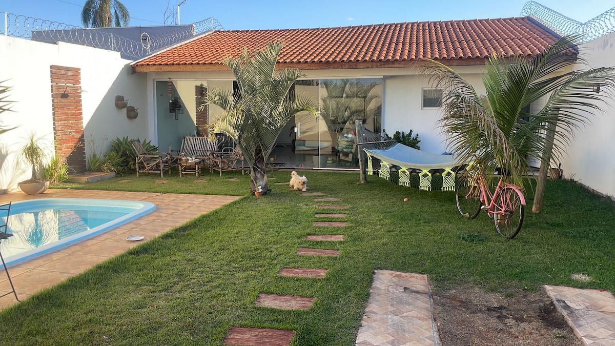 A spacious outdoor area features a swimming pool surrounded by lush green grass. A hammock is seen between two palm trees, while a pathway made of stepping stones leads toward seating arrangements and a bicycle nearby. The exterior of the main house is visible in the background.