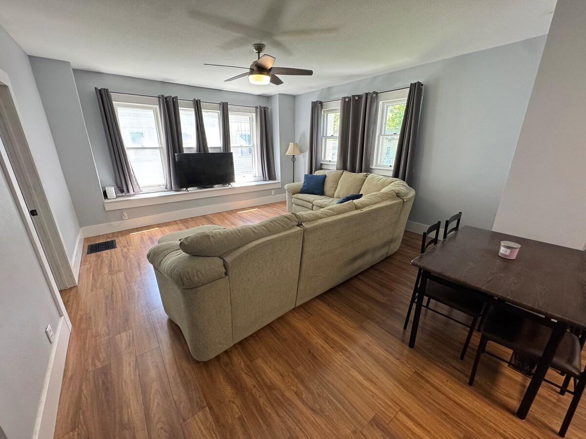 A spacious living area is visible, featuring large windows with sheer curtains that allow natural light to enter. A comfortable light-colored sectional sofa is positioned near a TV, and a small dining table with four chairs sits in the corner, enhancing the inviting space.