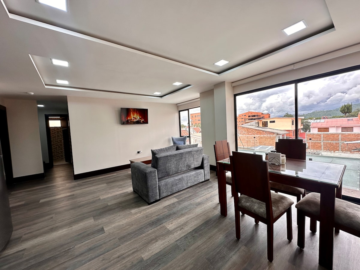 An open living area is showcased with large windows providing natural light. A sleek dining table with chairs is positioned beside a comfortable sofa, while a wall-mounted television adds functionality. The modern decor features wood-like flooring, enhancing the spacious environment.