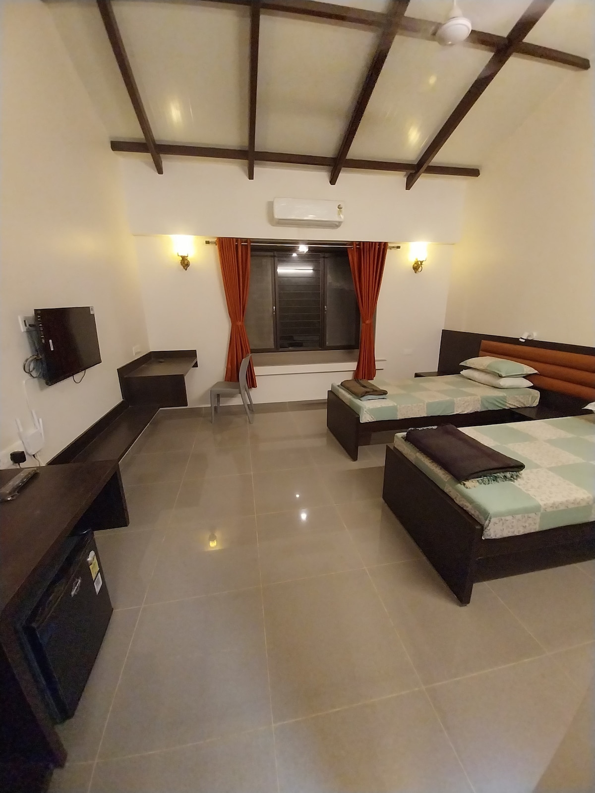 The room features two spacious beds with a quilted design. A flat-screen television is mounted on the wall opposite a sleek desk and chair. Natural light filters through a large window, which is augmented by orange curtains. A mini-fridge is present, providing added convenience.