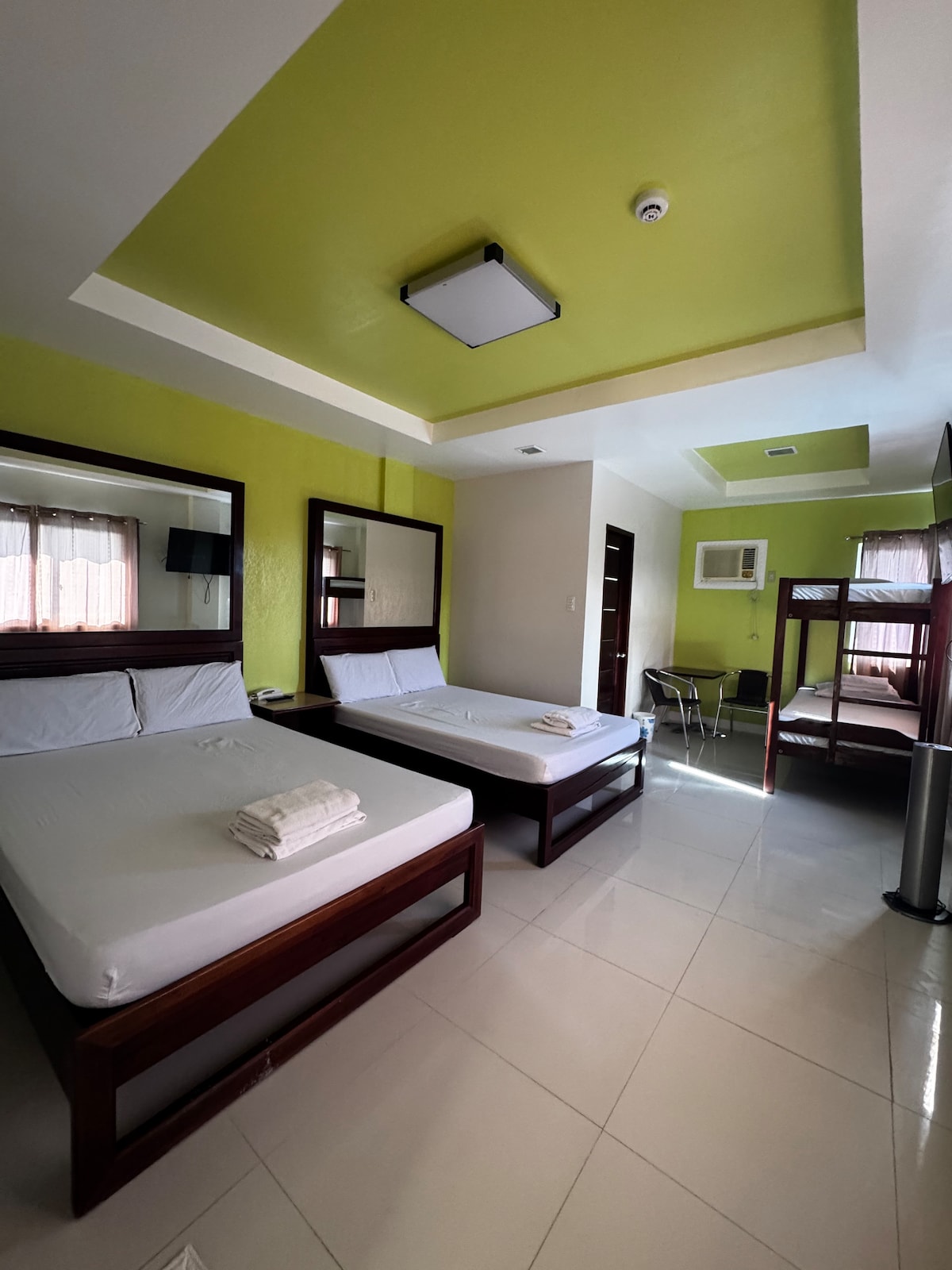 A well-lit room features two beds with crisp white linens and wooden frames. Mirrors are positioned above each bed, reflecting the vibrant green walls. A small table and chairs are situated in one corner, and light is provided by a ceiling fixture.