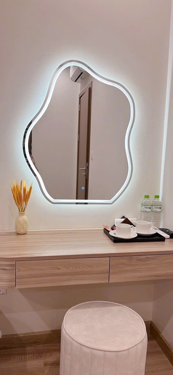 A uniquely shaped mirror is illuminated along its contours, reflecting the light from the room. A wooden vanity is positioned beneath it, accompanied by a minimalistic stool. A small vase with dried wheat sits alongside two bottles of water and a tray holding two cups.