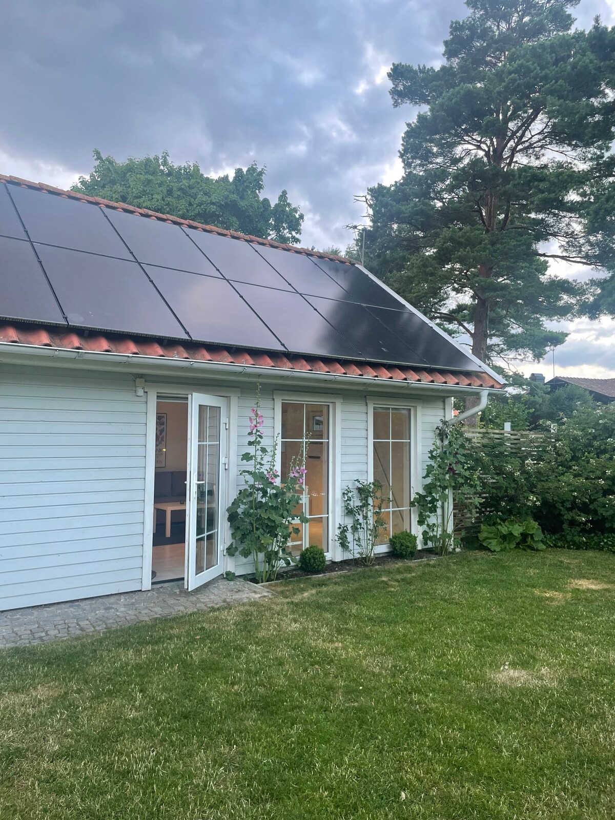 A modern guesthouse is presented, featuring a light blue exterior with solar panels on the roof. Tall windows flanked by climbing plants allow natural light to enter, while a well-maintained grassy area surrounds the building, providing a serene outdoor space.
