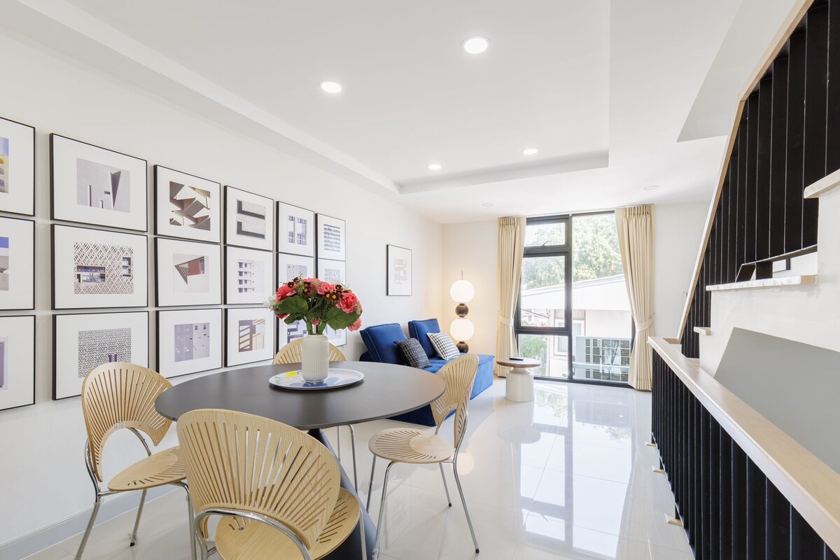 A bright living area features a round dining table surrounded by four wooden chairs. A blue sofa is positioned against a wall adorned with framed artwork. Natural light streams through a large window, highlighting the polished floors and modern decor.