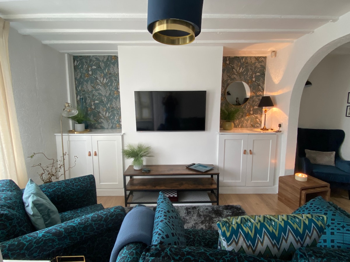 The living room features a modern design with a large flat-screen television mounted on a white wall. Two stylish sofas in deep teal are positioned around a wooden coffee table. Decorative elements include a floor lamp, a round mirror, and a touch of greenery.