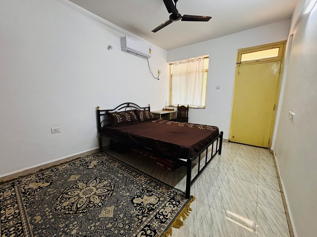 A spacious bedroom is presented with a dark bed spread covering a large bed, complemented by a patterned rug on the floor. A window allows natural light to filter in through sheer curtains, and a ceiling fan is mounted above, providing airflow.