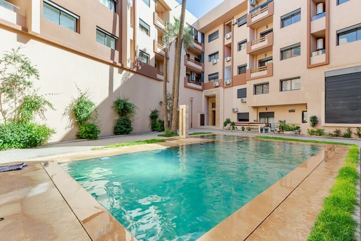 A serene private pool is surrounded by lush greenery and light-colored stone paving. The building facade features multiple windows and balconies, allowing natural light to enhance the inviting ambiance of the outdoor space.