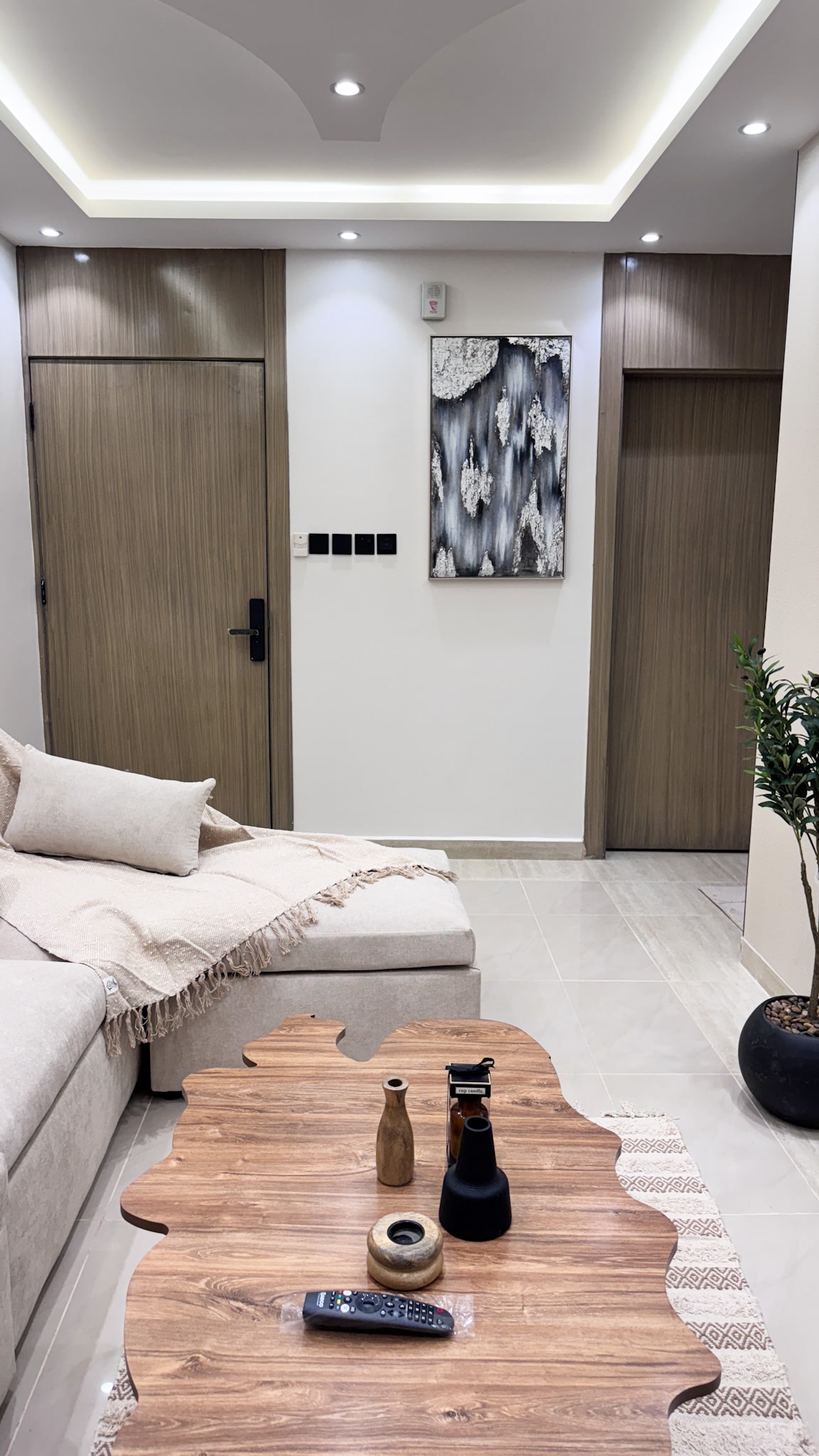 A modern living area is presented with a light-colored sofa adorned with a neutral-toned throw. A wooden coffee table with a unique shape is centered in front, featuring decorative items. Two doors are positioned on the sides, and a small potted plant adds a touch of greenery.