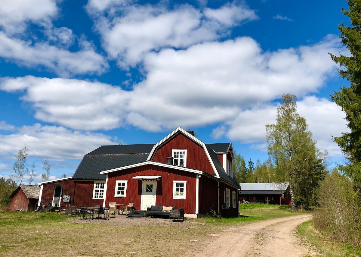 The charming red house features a mix of traditional and modern architectural elements, set against a backdrop of a clear blue sky dotted with fluffy white clouds. A dirt path leads up to the house, surrounded by greenery and distant trees.