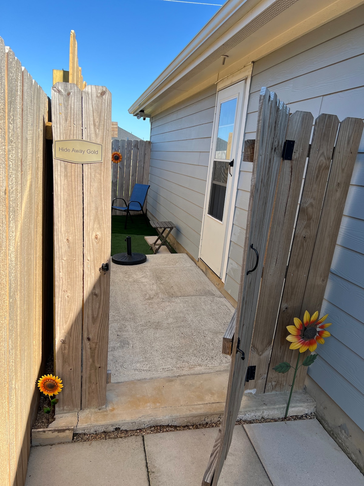 A private entryway opens to a small patio area, featuring a chair and a small table. The entrance is framed by a wooden gate with decorative sunflowers, and a door leads into the guesthouse. Clear blue skies are visible above.