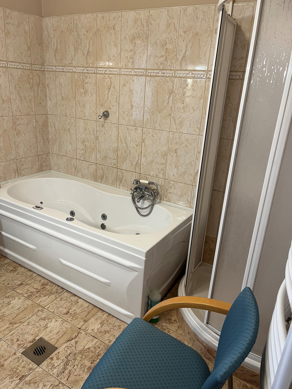 The bathroom features a white jetted bathtub alongside a clear glass shower stall. Light-colored tiles surround both fixtures, with a small drain visible in the floor. A blue upholstered chair is positioned nearby, adding a functional touch to the space.