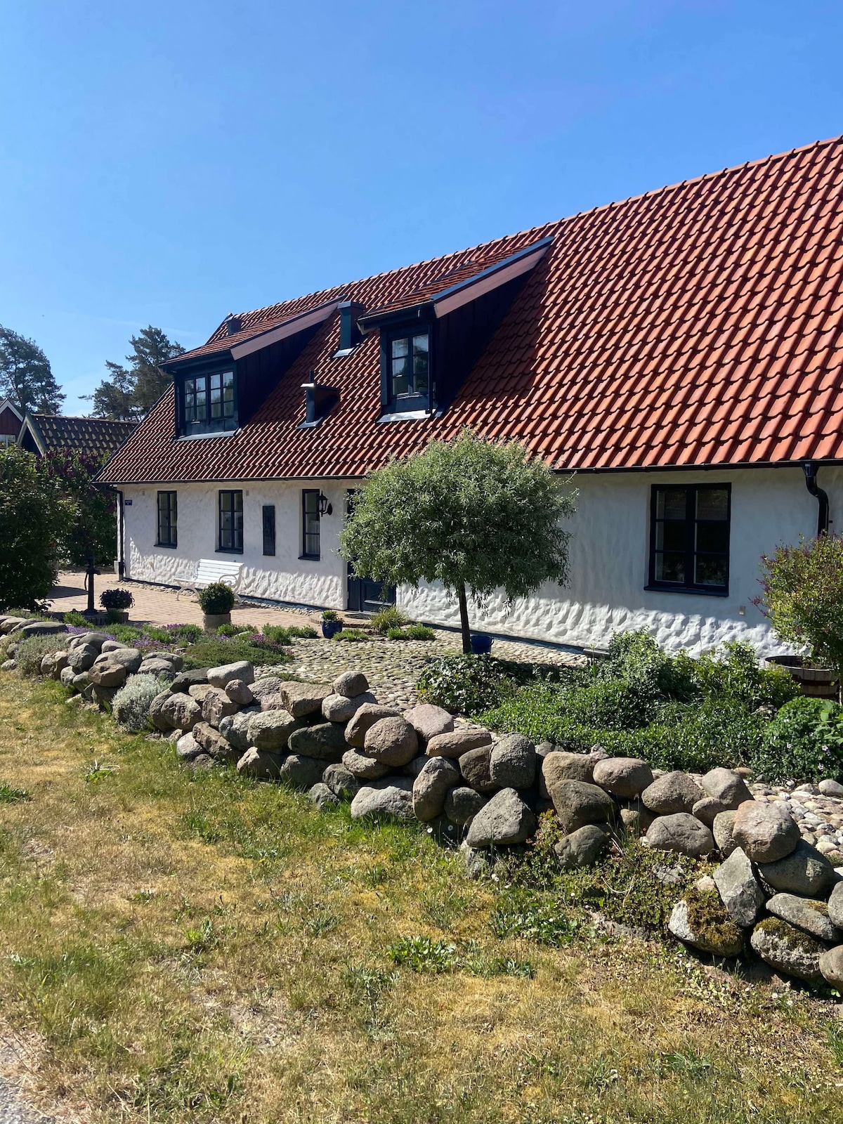 The exterior of the house features a classic design with a red-tiled roof and white walls. A well-maintained garden is visible, surrounded by a stone wall. Several windows allow natural light to enter, and a small tree adds greenery to the inviting façade.