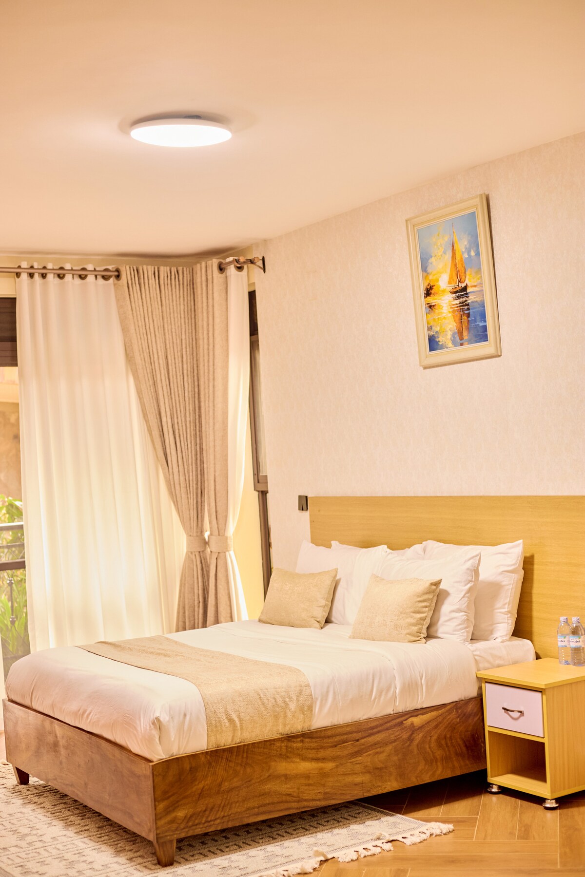 A restful bedroom is shown with a spacious bed dressed in white linens and accented with decorative pillows. Natural light filters through sheer curtains, and a warm-toned wooden nightstand sits beside the bed, featuring a glass of water. A painting of sailboats adorns the wall.