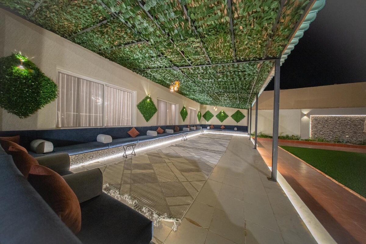 A spacious outdoor lounge area features a partially shaded seating arrangement beneath a canopy of greenery. Soft cushions are placed on a textured surface, while ambient lighting runs along the perimeter, creating a calm atmosphere. Lush landscaping is visible in the background.