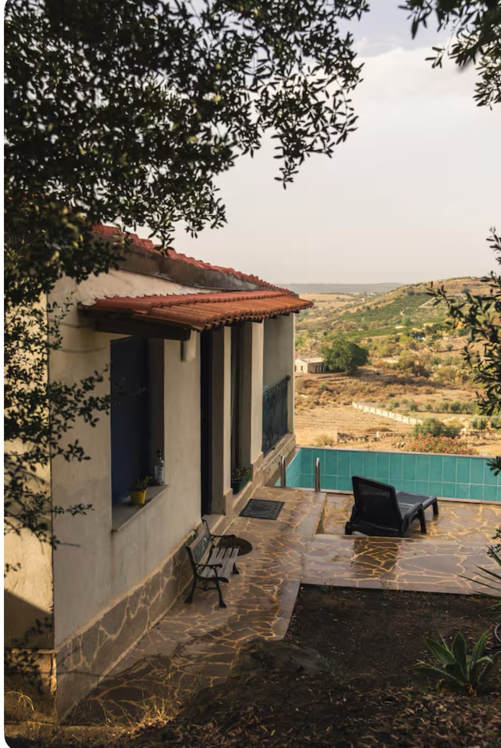 A scenic view of the house features rustic architecture with a terracotta roof. A chaise lounge rests on the stone patio, surrounded by greenery. The private pool glimmers in the background, framed by rolling hills and valleys extending into the distance.
