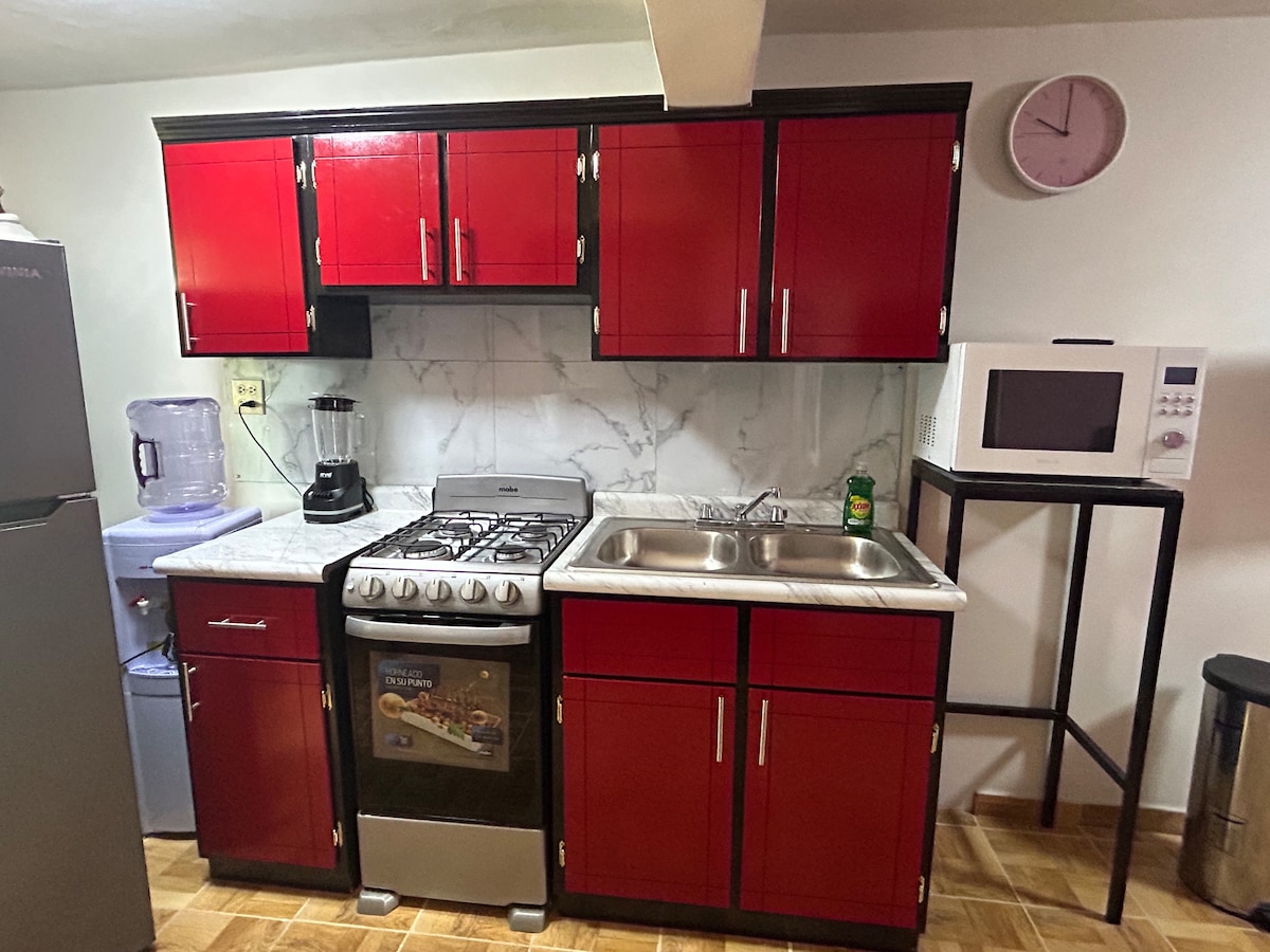 A modern kitchen setup features red cabinetry with a marble backsplash. Stainless steel appliances include a gas stove, microwave, and a blender. A kitchen sink is positioned between the stove and a compact refrigerator, while a wall clock is mounted above for convenience.