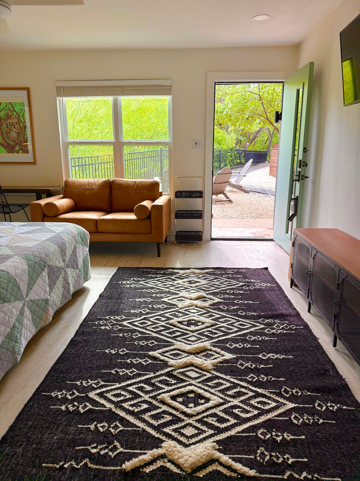 A cozy bedroom features a spacious bed with a patterned quilt and a stylish armchair in warm tones. Natural light filters through large windows, illuminating a textured area rug. A door leads to an outdoor space, enhancing the connection with nature.