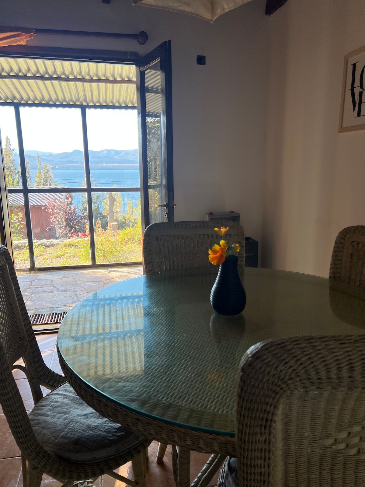 A dining area is presented with a round glass table surrounded by four woven chairs. A black vase containing vibrant flowers is centered on the table. In the background, large glass doors open to reveal a scenic view of the lake and distant mountains.