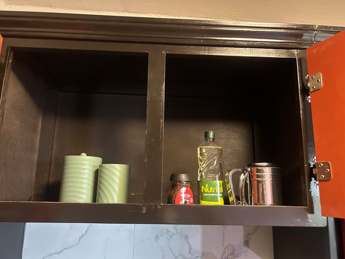 An upper kitchen cabinet displays two green containers with ribbed surfaces, a jar of instant coffee, a bottle of cooking oil, and a metal cup. The cabinet's doors are painted in a warm brown hue, providing a subtle contrast to the light-colored background.