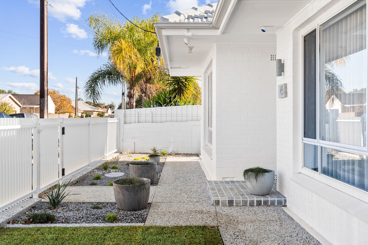 A clean, modern entrance is showcased with a white brick exterior and well-maintained landscaping. Paved pathways lead to the front door, flanked by decorative pots and textured gravel. Lush greenery and a white picket fence enhance the inviting outdoor space.
