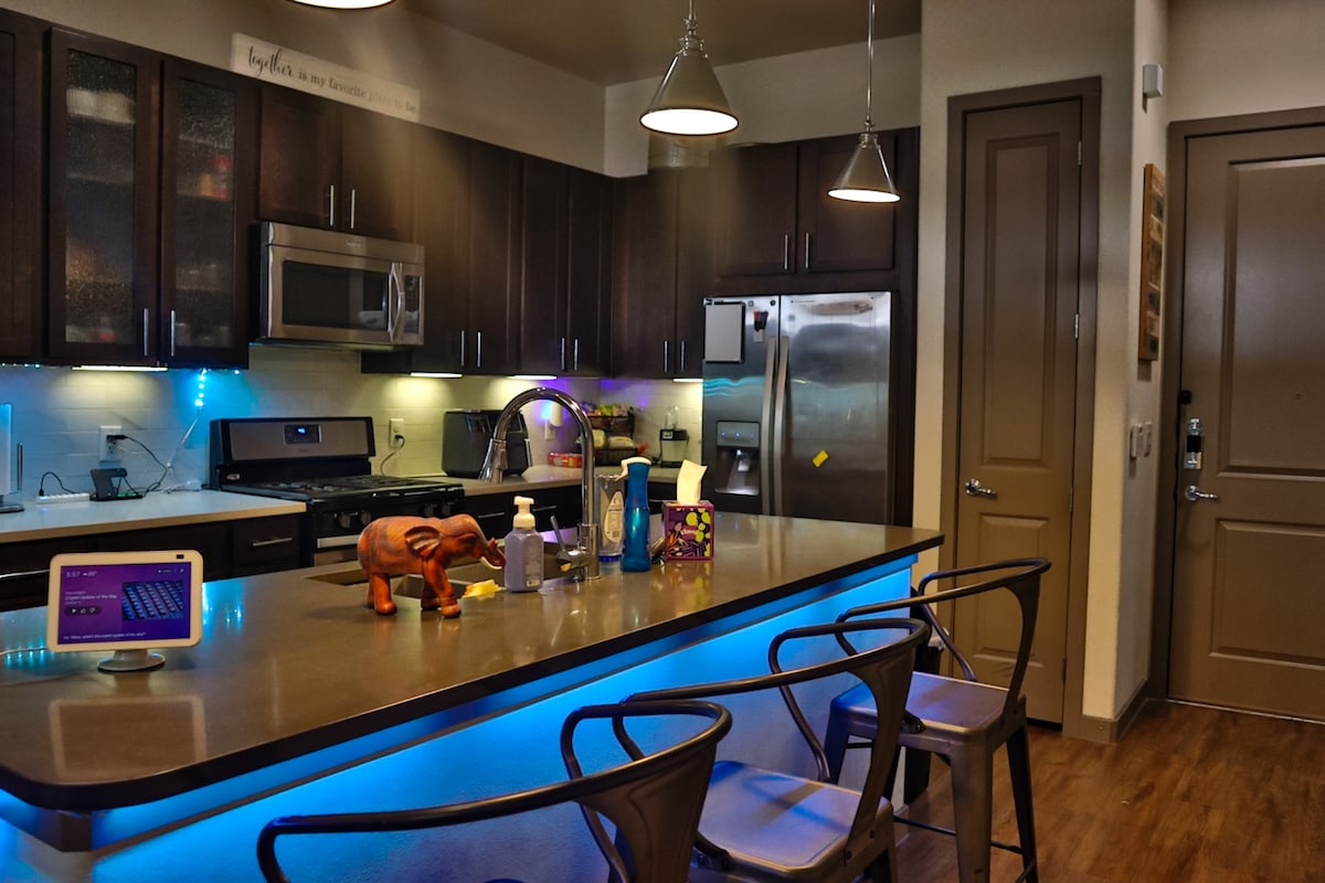 A modern kitchen is displayed, featuring stainless steel appliances and a large countertop. Under-cabinet lighting casts a soft glow. Several bar stools are arranged around the island, complemented by decorative items and cleaning supplies on the counter. The entrance door is visible, enhancing the open design.