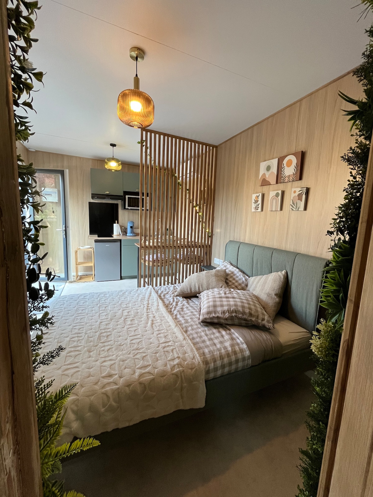 A cozy sleeping area is divided by a wooden screen, creating a sense of privacy. A spacious bed is topped with neutral bedding and decorative pillows. The room features light wood paneling, framed wall art, and glimpses of kitchen amenities in the background.
