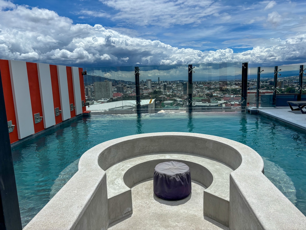 A rooftop pool area features a circular seating arrangement beside the water, with a plush cushion in the center. The skyline of the city is visible in the background, framed by a dramatic sky and large glass panels providing safety and views.