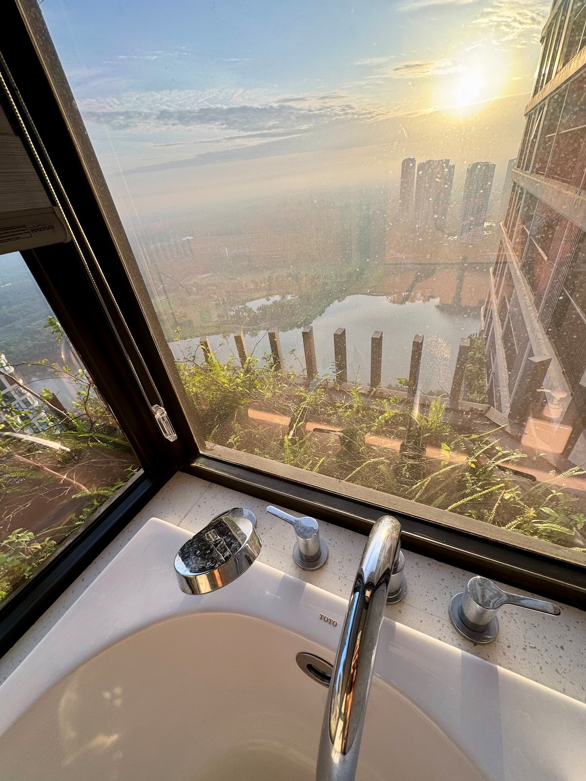 A modern bathtub is positioned by a large window, offering a sweeping view of a serene landscape with the sun rising above distant buildings. Fresh greenery is visible outside, enhancing the luxurious ambiance of the space.