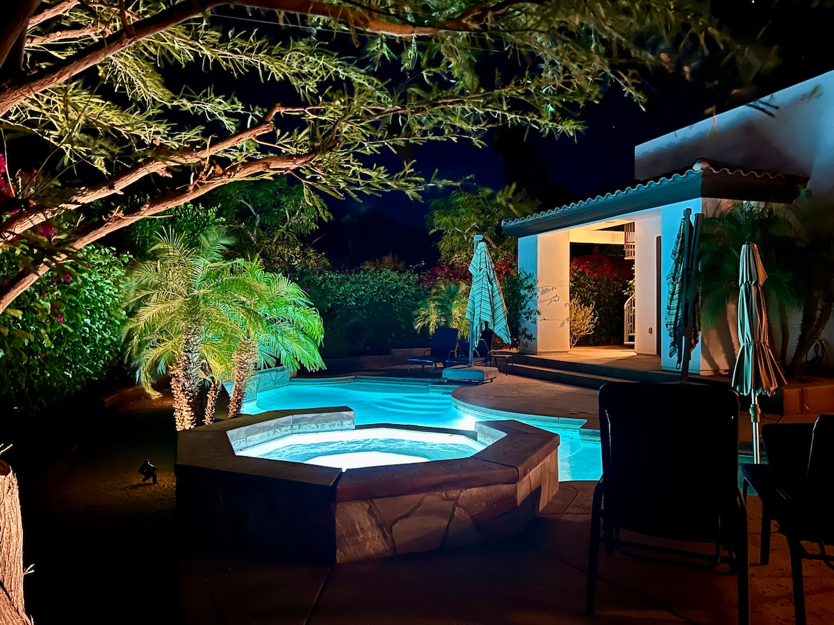 A serene outdoor space is illuminated at night, featuring a private hot tub surrounded by lush greenery and palm trees. Lounge chairs and umbrellas are visible nearby, with a softly lit patio area enhancing the tranquil environment.