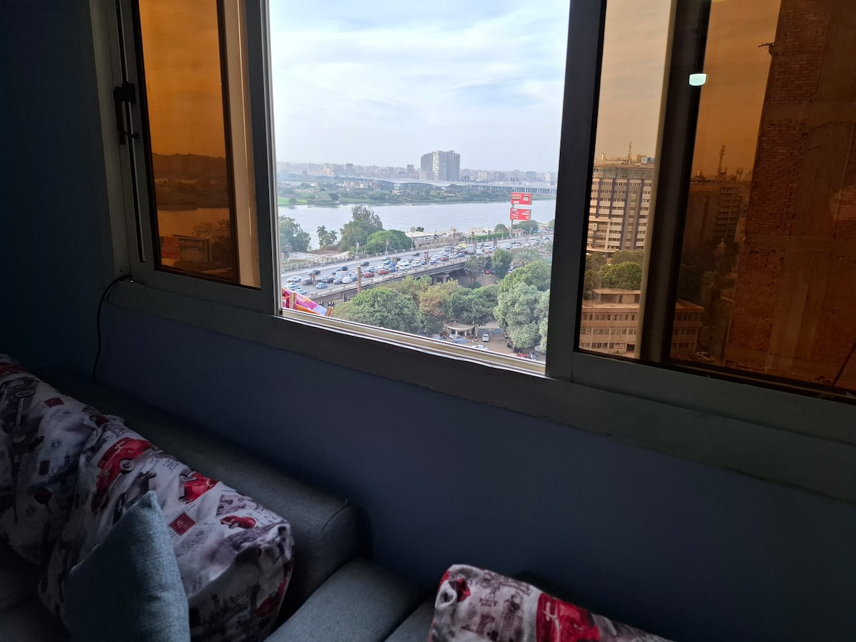 A view of the Nile River is captured through large windows, showcasing a bustling cityscape. Soft gray sofas are positioned nearby, with patterns visible on the cushions. The warm light filters in, adding a cozy atmosphere to the space.