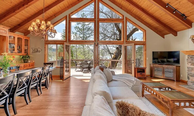 The open living area features soaring wood ceilings and large glass doors, allowing natural light to fill the space. A comfortable sectional sofa is positioned near a stone fireplace, with a dining area visible in the background. Views of the outdoor deck and surrounding trees can be seen.