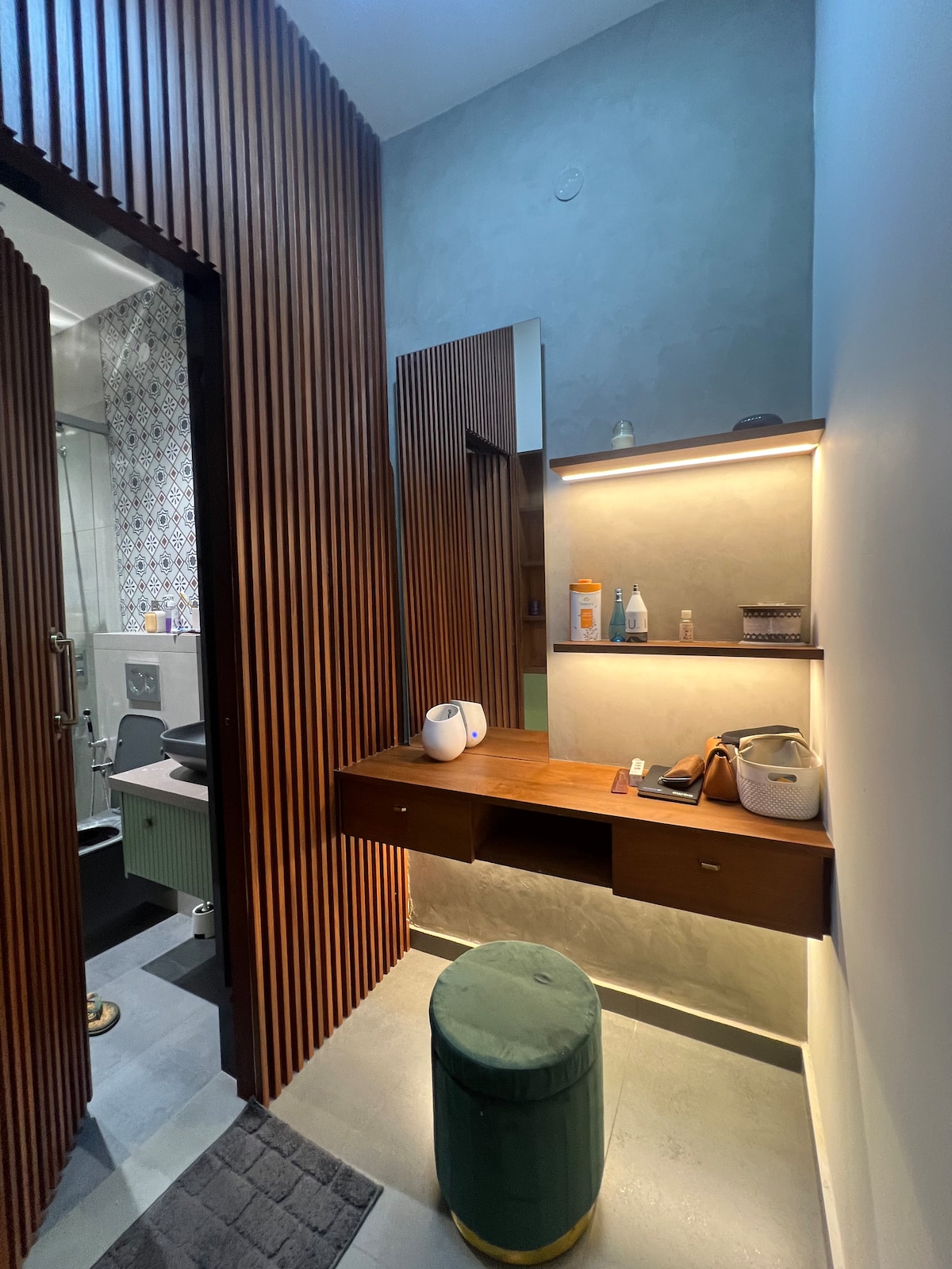 The bathroom area features wooden paneling and a sleek vanity with a large mirror above it. A small green stool is positioned in front of a wooden counter, which displays toiletries and small items. A shower area is partially visible through an open door.