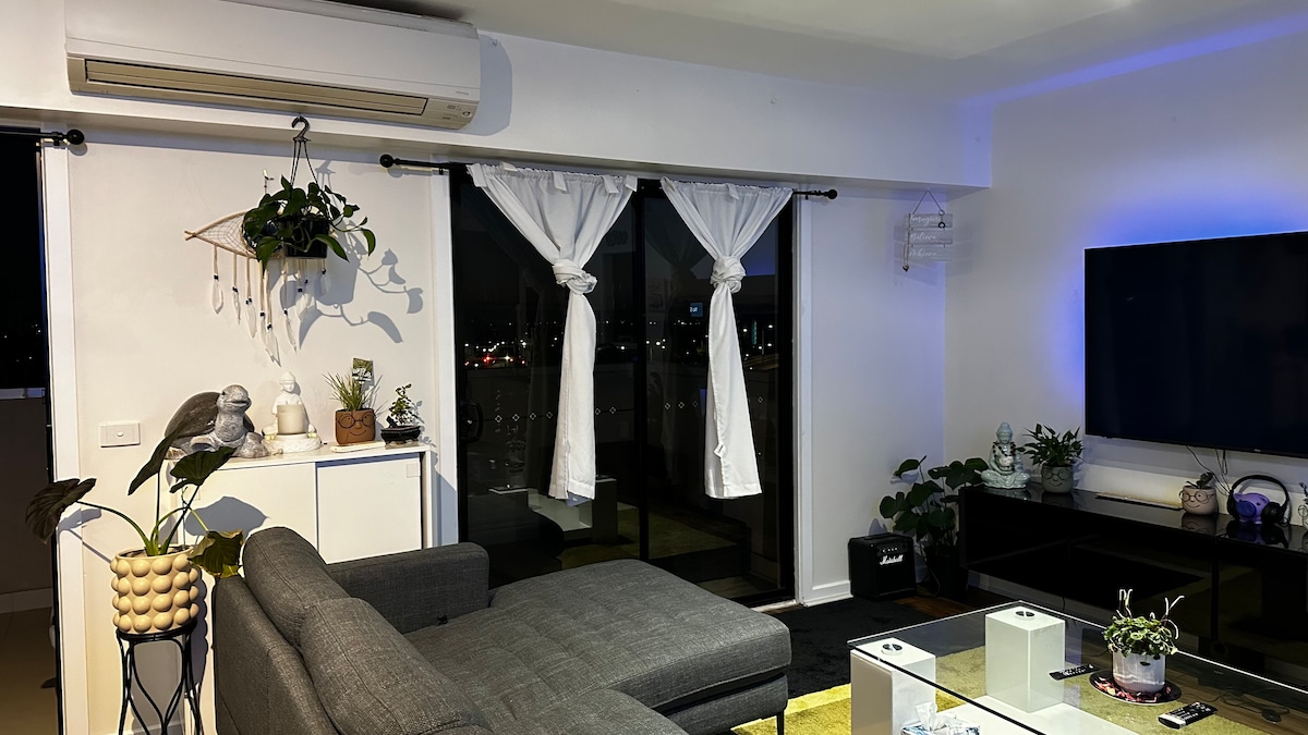 A living area is presented with a modern gray sectional sofa and a glass-top coffee table. Bright natural light enters through large balcony doors, covered by sheer white curtains. A flat-screen television is mounted on a dark entertainment unit, while various potted plants add greenery to the space.