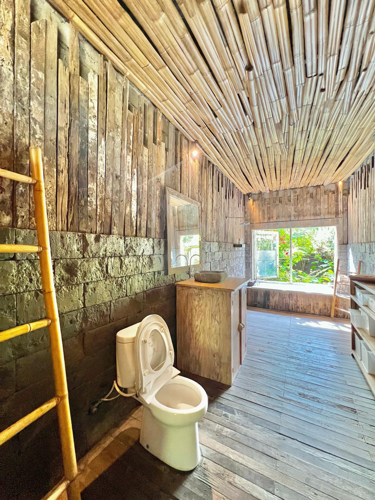 The bathroom features rustic bamboo walls and a wooden vanity, providing a natural aesthetic. Natural light pours in from the window, illuminating the space. A contemporary toilet is positioned beside a ladder rack, enhancing the functional layout.
