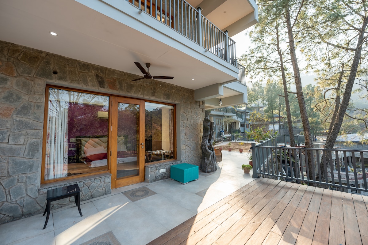 An outdoor patio area is showcased with a wooden deck and stone walls. A small black chair is positioned near glass doors, leading into the interior. Complementary blue seating adds a pop of color, while surrounding greenery and a sculpture provide an inviting atmosphere.