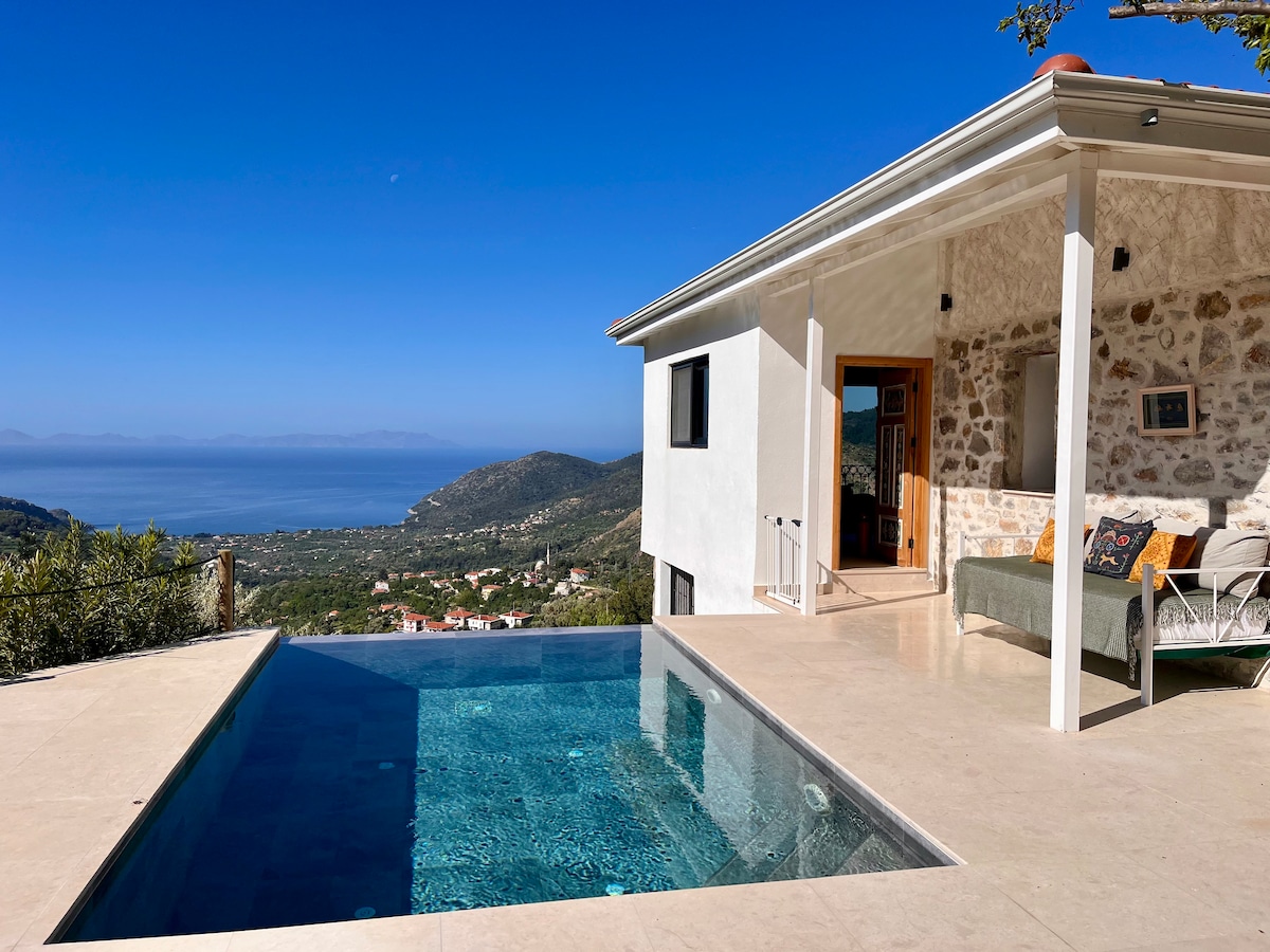A serene outdoor space features a clear infinity pool, offering sweeping views of the landscape and sea. The house displays a mix of natural stone and modern architecture, with a covered patio area that includes comfortable seating arrangements.