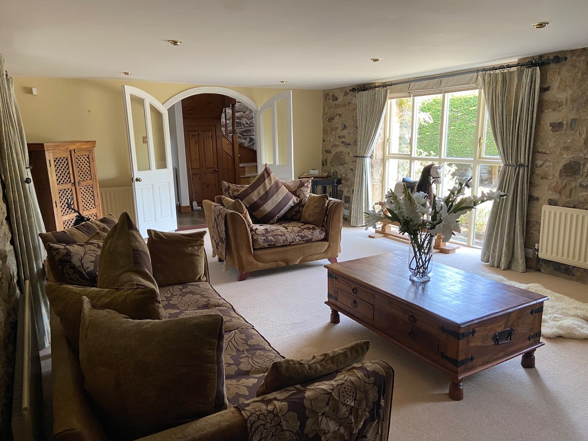 A comfortable living area showcases plush sofas arranged around a wooden coffee table. Soft lighting from large windows highlights the textured stone walls. Decorative elements, such as flowers on the table and patterned cushions, add subtle charm to the inviting space.