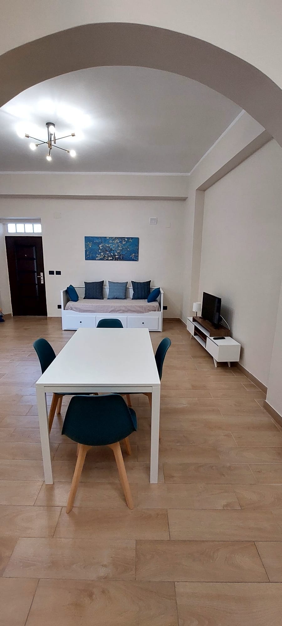 An airy living space features a white dining table surrounded by teal chairs. A comfortable seating area with decorative cushions is visible against the wall. The room is illuminated by overhead lighting, and a television is positioned on a sleek entertainment unit.