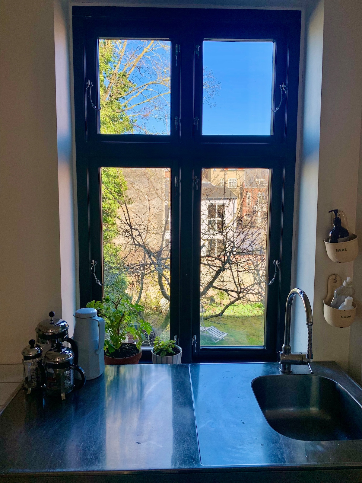 A stainless steel sink is positioned below a large window, allowing natural light to illuminate the space. A coffee maker and a teapot are displayed on the counter alongside small potted plants. Outside, trees and a garden view are visible, creating a serene backdrop.
