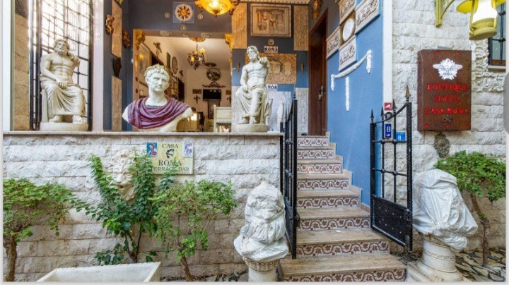 An elegant entrance features intricately designed stone steps leading to the lobby. Marble statues of historical figures are positioned on either side, adding a classic touch. Lush greenery lines the pathway, complemented by decorative tiles that enhance the welcoming ambiance.