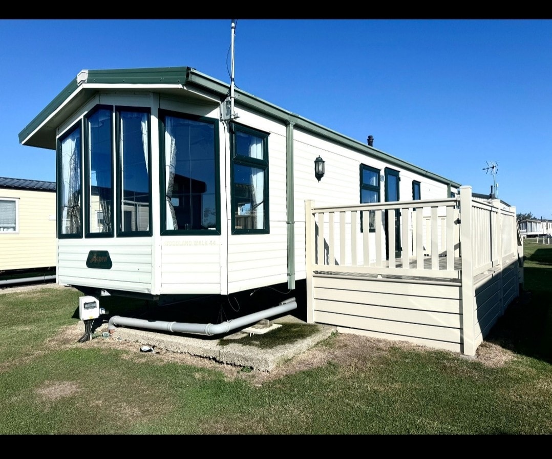 A stylish caravan situated on a grassy area features large front windows, allowing natural light to enhance the open living space. A private deck wraps around the entrance, providing outdoor accessibility. The exterior displays a clean, neutral color palette, complementing the surrounding landscape.
