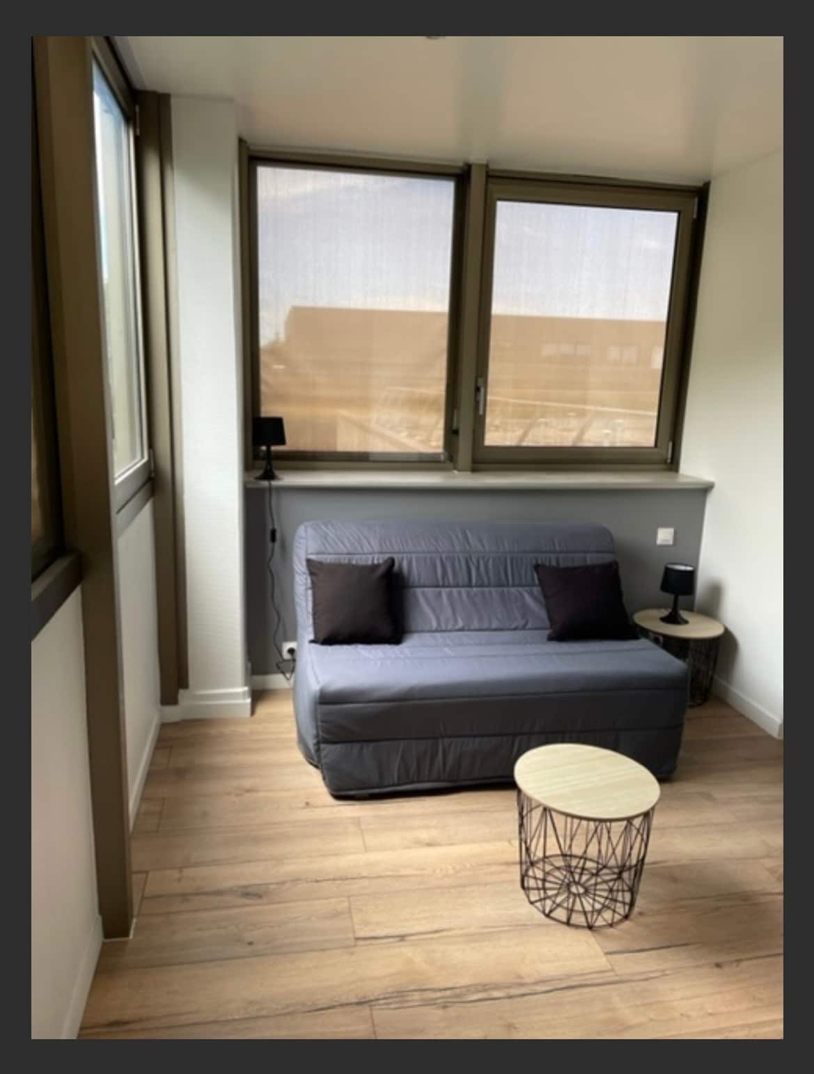 A compact seating area is presented with a gray sofa that converts into a bed, adorned with two black cushions. A small round table with a wire design is positioned beside the sofa. Natural light filters through two partially opened windows, enhancing the space.