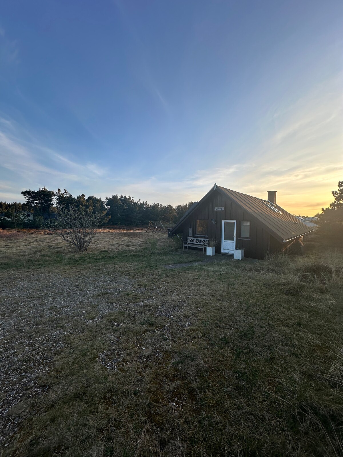 A charming summer house is set against a backdrop of trees and open fields. The structure features an angled roof and a welcoming entrance. Gray gravel paths lead up to the house, and the expansive grassy area surrounding it captures the natural beauty of the serene landscape.