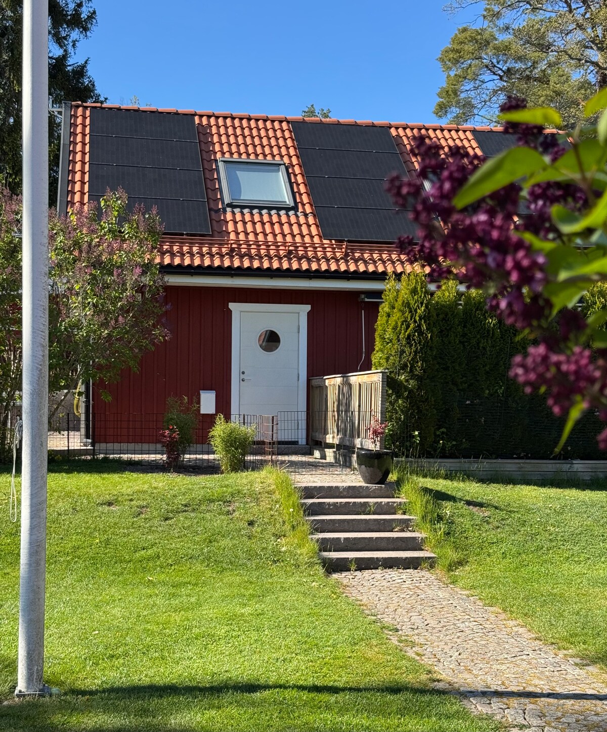 A charming house with a red exterior and a tiled roof is showcased. A front door features a circular window, while solar panels are mounted on the roof. A pathway lined with stone leads up to the house, surrounded by green grass and shrubs.