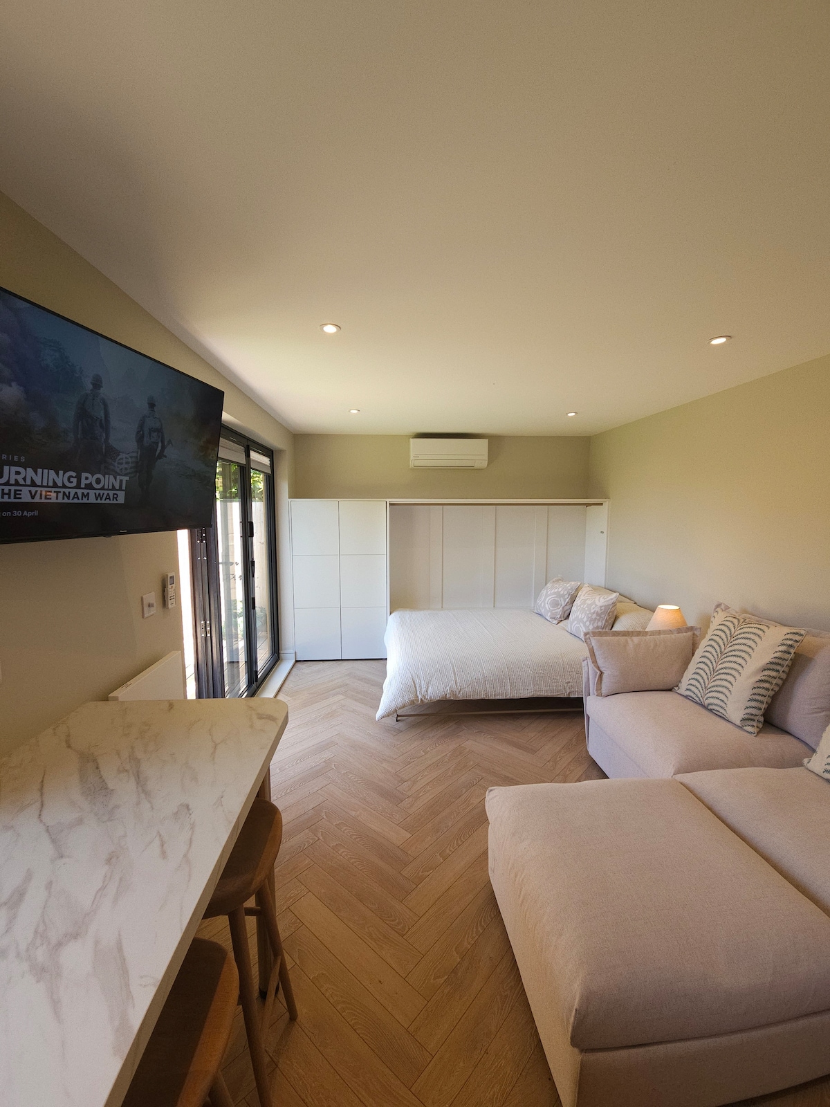 The studio apartment features a modern design with light-colored walls and a herringbone-patterned floor. A spacious sofa and a fold-down bed are visible, along with a mounted television. Light enters through bi-fold doors that lead to the private garden area.