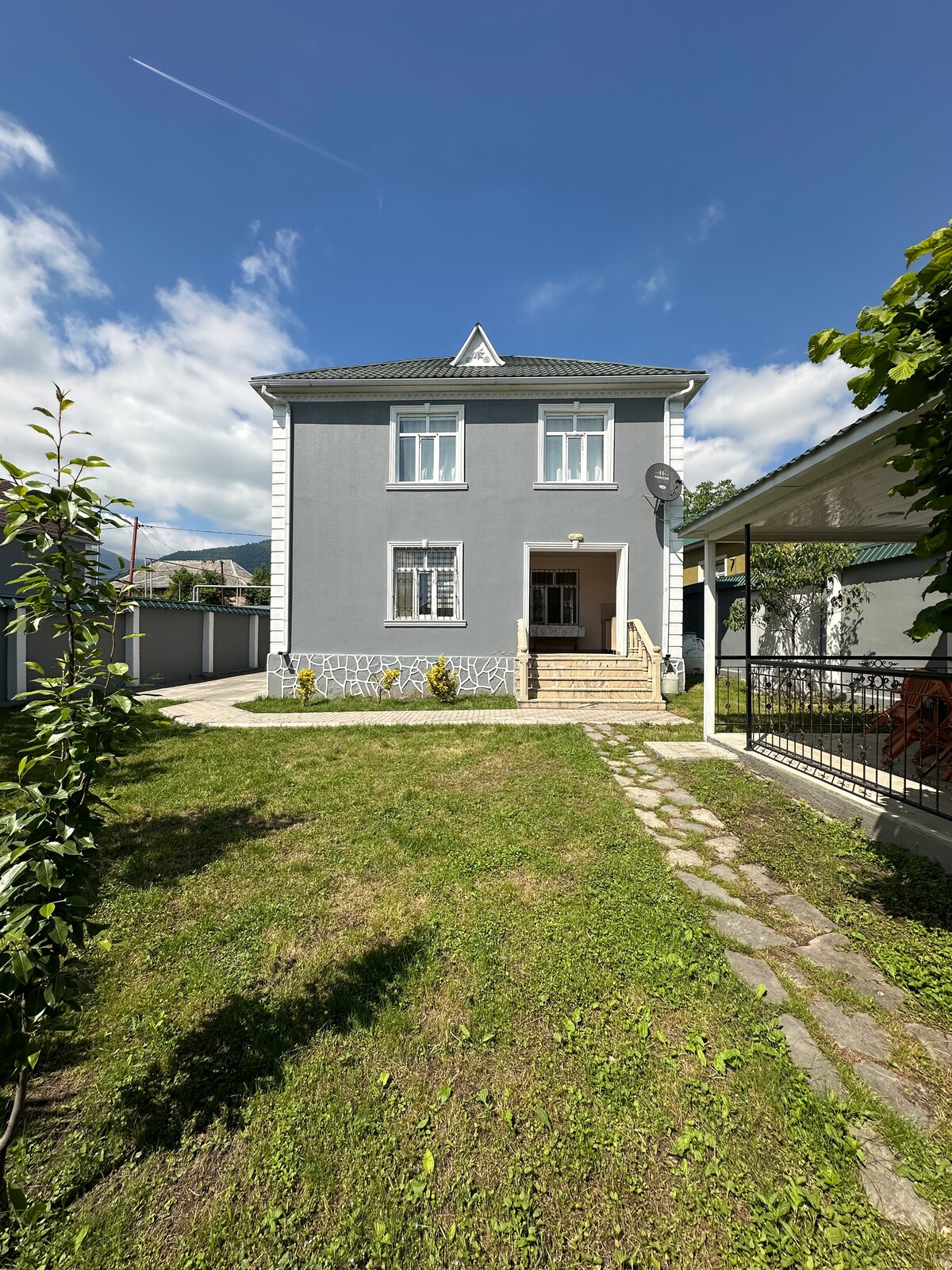 A two-story house is presented with a light gray exterior and a well-maintained lawn. The entrance features a set of steps leading to a welcoming door, while a paved pathway is highlighted by two rows of greenery on either side. Clear blue skies complement the scene.
