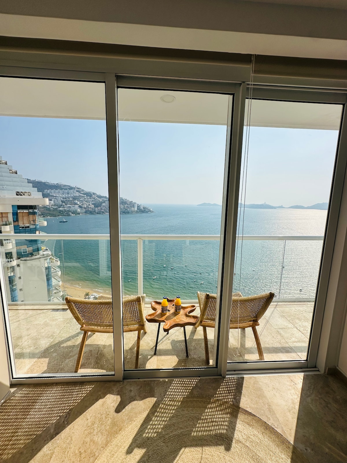 A small balcony is visible with two woven chairs facing the ocean. A round table is set between them, featuring glasses and a pitcher, all placed against a backdrop of tranquil waters and distant hills. Floor-to-ceiling glass doors frame the stunning view.