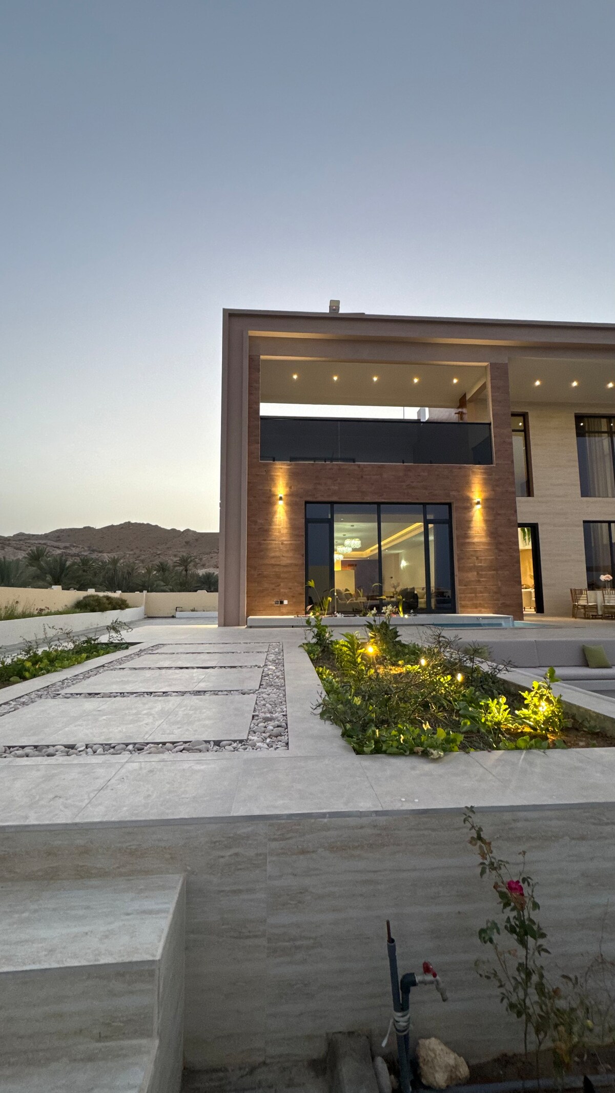 A modern villa exterior is showcased, featuring large glass windows and elegant stone accents. Well-manicured landscaping details pathways lined with greenery and flowers. Soft lighting highlights the architectural elements, creating a serene atmosphere against the backdrop of a fading sunset.